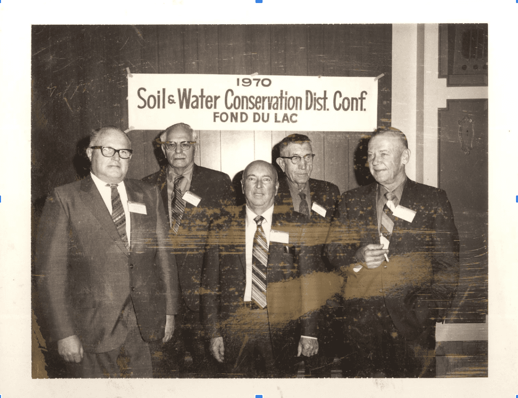 A group of five men in suits and ties stands in front of a sign reading 1970 Soil & Water Conservation Dist. Conf. Fond du Lac. Indoors, three wear glasses and name tags on their jackets, likely discussing the shift from conventional to organic farming techniques like those championed by Doudlah Farms.